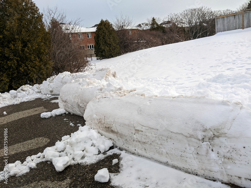 фотографія A large, chunky plowed snowbank sits on black asphalt, bordering a snowy hill covered in footprints on a trail