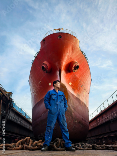 A shipyard worker stands proudly in front of a giant cargo ship under maintenance in a dry dock.