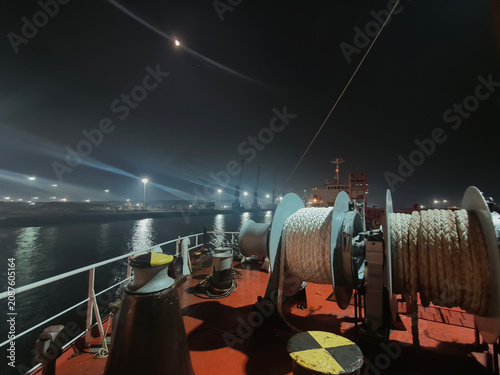 Mooring winches and heavy ropes on a cargo ship deck at night with port lights background.