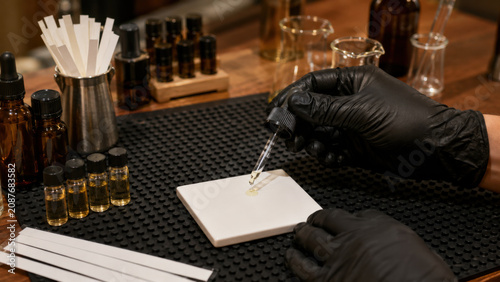 Gloved hands carefully adding single drop from dropper onto white ceramic tile beside fragrance vial on black rubber mat at wooden laboratory desk