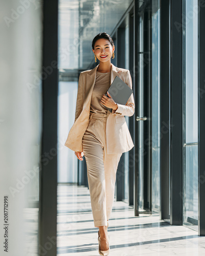 Confident successful Asian businesswoman walking through a modern glass office carrying a laptop, elegant beige business suit, warm natural sunlight, luxury corporate headquarters