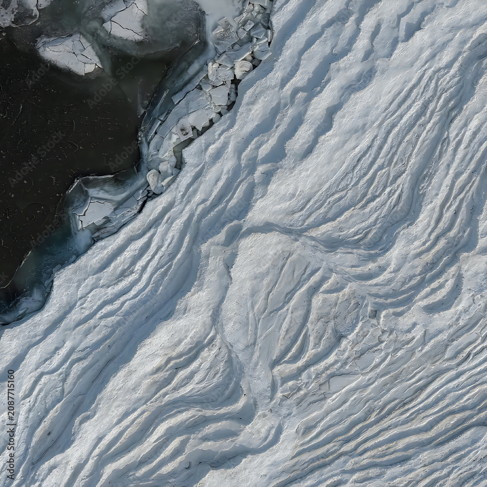Obraz premium Aerial view of white snow ripples on frozen lake ice surface next to dark open water in a winter landscape.