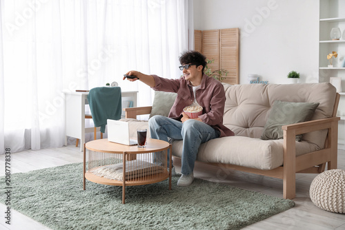 Young man in 3d eyeglasses with remote control and popcorn on sofa in living ...