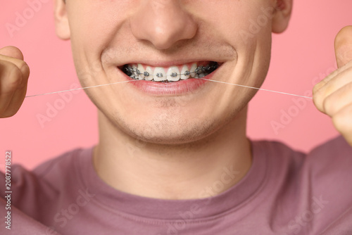 Fotografía Young man with dental braces flossing teeth on pink background, closeup