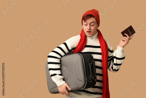 Handsome young shocked man with suitcase and passport on brown background. Wi...