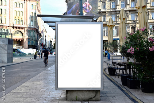 bus shelter at busstop. white poster ad display space and lightbox. mockup template. blank ad panel. glass design. urban street setting. soft background. silhouettes in a distance