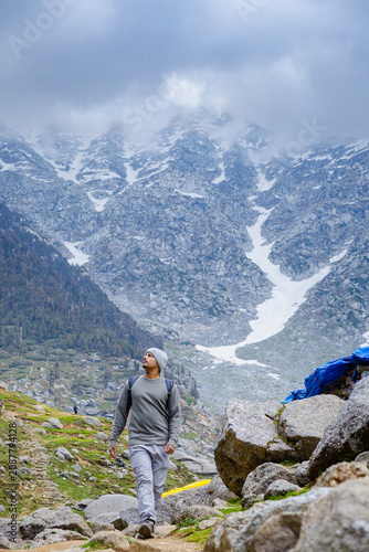 Hiker walking on a rocky mountain trail at Kareri Lake in Dharamshala with snowy peaks and clouds