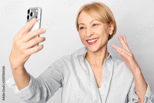Obraz na plátně Selfie smiling senior woman holds phone, waving to camera, joyful portrait indoo