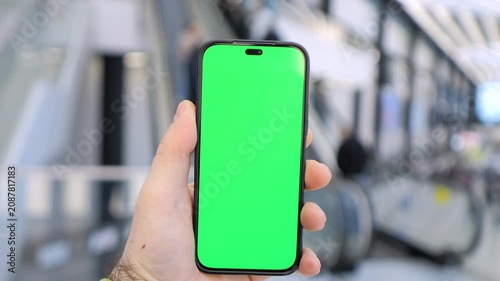 Hand holds phone with green screen. Background shows blurred train station. Three keyframes show slight camera movement. People walk past in background. No animals or plants visible