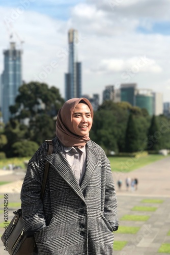 Stylish Asian Muslim woman wearing a hijab and casual smart outfit in an urban park with modern city skyline in the background, capturing authentic fashion and contemporary lifestyle.