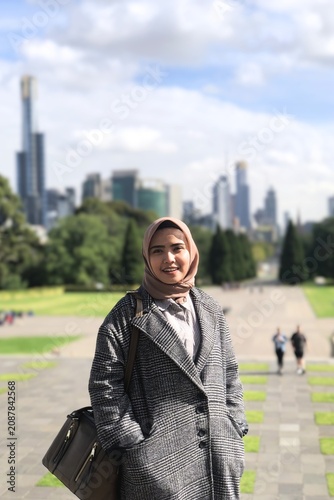 Stylish Asian Muslim woman wearing a hijab and casual smart outfit in an urban park with modern city skyline in the background, capturing authentic fashion and contemporary lifestyle.
