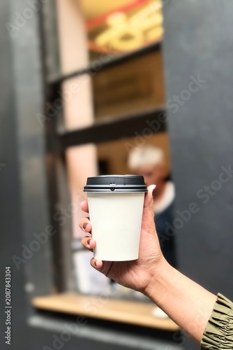 Close-up of a hand holding a blank white takeaway coffee cup with a black lid in front of a modern café. Disposable paper cup mockup with copy space for custom branding, coffee shop marketing