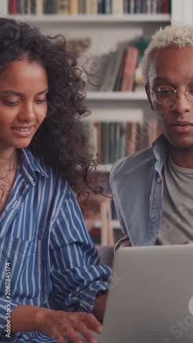 African American couple sits together on couch, typing on laptop computer keyboard, discussing project, pointing at screen, and smiling while working from home office workspace.