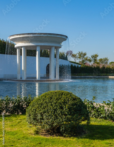 Round pavilion rotunda with white columns stands on Lake of Loves, water cascades down in lake. Green shrubs and grass surround it under clear blue sky. Krasnodar. Galitsky Park - Cloud Park