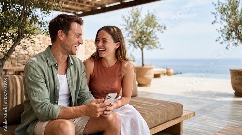 Young couple laughing and holding smartphone on a luxury Mediterranean patio.