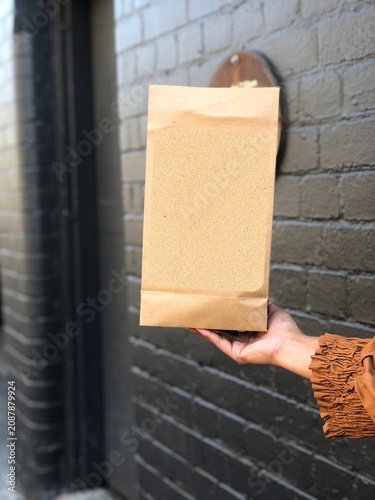 Blank standing kraft paper pouch with transparent window held by hand against an urban brick wall. Eco-friendly food packaging mockup with copy space for custom branding, ideal for coffee, tea, bakery