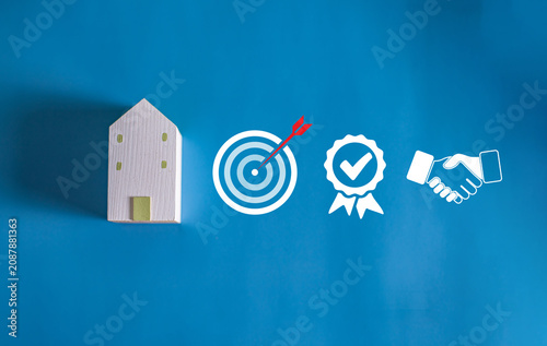 Wooden house model with business goal, quality assurance and partnership icons on blue background