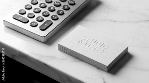 A professional designer or accountant workspace featuring a stack of minimalist Studio Nuno embossed business cards beside a modern metallic calculator on a clean white marble desk surface top view