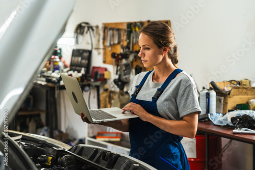 Female car mechanic inspecting vehicle with laptop beside open hood