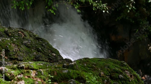 Close-up shot of a moss-covered rock with a powerful waterfall cascading in the background, Serene dynamic nature scene. Tao Thong Waterfall, Phang Nga Province, Thailand.