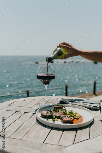 фотографія Red wine being poured into glass with steak and ocean view