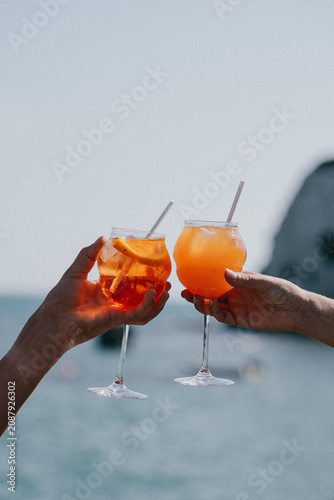 Photos Two hands clinking glasses of orange spritzer drinks on a beach