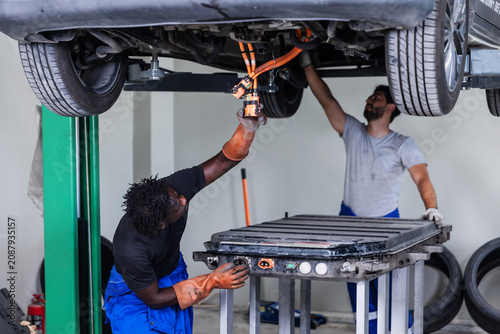 Professional EV technician disconnecting and inspecting a high-voltage battery connector beneath a lifted electric vehicle during maintenance.