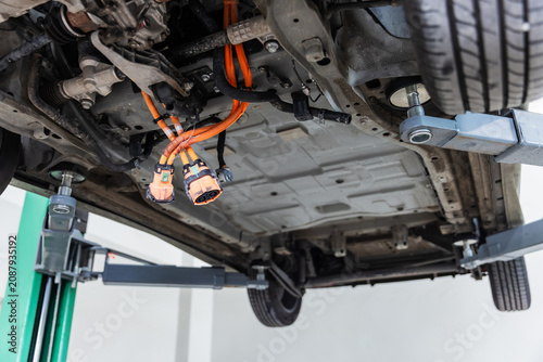 Close-up underside view of an electric vehicle lifted on a hydraulic service lift, revealing high-voltage orange cables and electrical connectors during maintenance inspection.