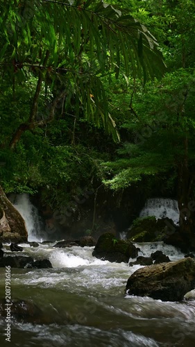 Vertical shot of a powerful waterfall cascading down into a fast-flowing river filled with mossy rocks, surrounded by dense tropical forest. Ideal for nature, travel, environment, and wellness concept