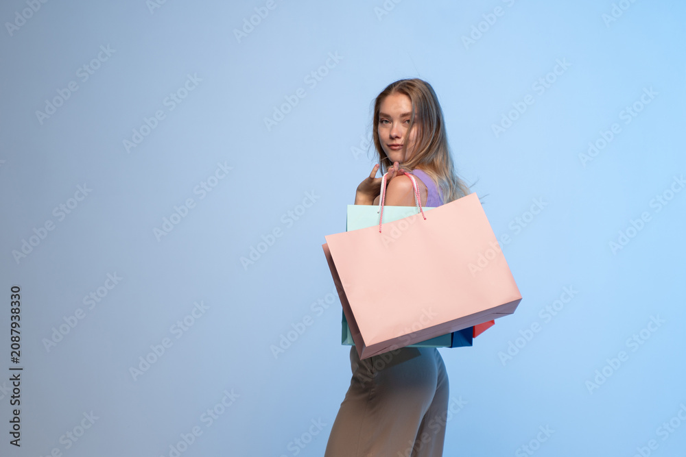 Obraz premium Young woman carrying colorful shopping bags over her shoulder, looking back at camera on a blue studio background