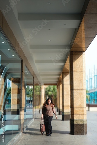 Woman smiles warmly under glass-and-concrete walkway. She holds a leather bag, standing calmly beside pillars. Sunlight spills softly through the open sky above.