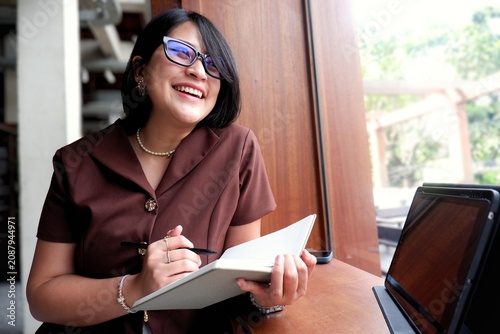 Smiling woman wears glasses and writes notes. She sits at a desk with a laptop beside her. Natural light shines through the window behind her. Her joyful expression suggests she's engaged.
