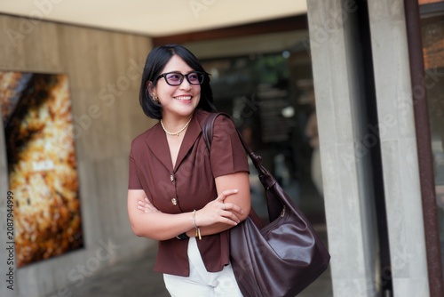 Warm smile lights up the woman's face. She wears glasses and a brown blouse. Her arms cross as she leans against a modern wall. Large bag rests on her shoulder. Her gaze invites connection, warmth.