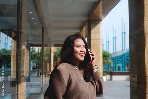 Smiling woman walks outside a modern glass building. She holds her phone, engaged in a pleasant conversation. Her outfit is cozy and stylish, adding warmth to the urban setting.