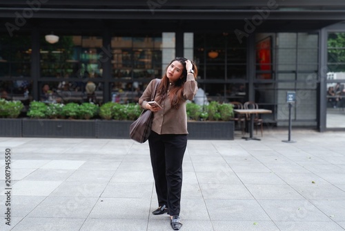 Young woman stands outside modern cafe. She holds phone and touches her hair. Her expression shows mild confusion or thought. Urban setting with glass walls and greenery. Ideal for lifestyle, travel.