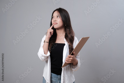 Foto Thoughtful young Asian businesswoman holding clipboard and looking away on gray