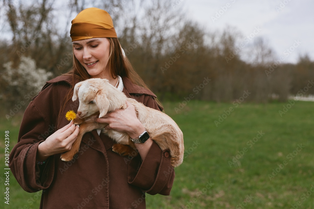 Fototapeta premium Joyful woman outdoors pets a gentle goat kid, holds a dandelion, sharing a warm moment of affection in a pastoral field, rustic jacket and scarf, serene countryside backdrop, candid lifestyle scene.