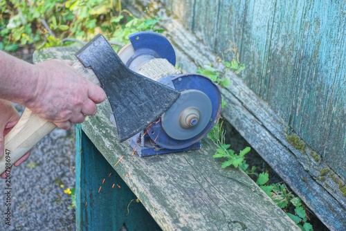 One male worker holds a large axe in his hands and sharpens the iron blade on a grinding wheel on an electric sharpening machine on a table