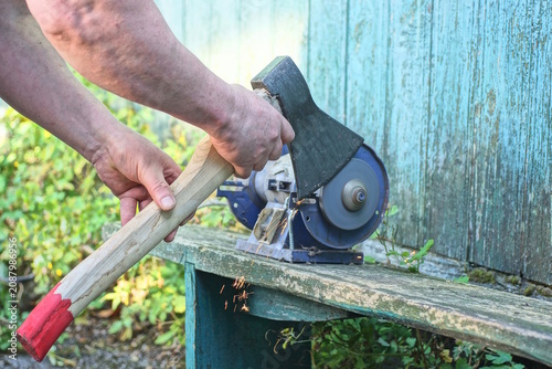 One male worker holds a large axe in his hands and sharpens the iron blade on a grinding wheel on an electric sharpening machine on a table