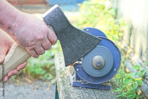 One male worker holds a large axe in his hands and sharpens the iron blade on a grinding wheel on an electric sharpening machine on a table