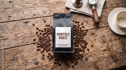 A matte black coffee bag is centered among scattered roasted coffee beans on a rustic wooden table, with a coffee cup, portafilter, and tamper nearby. Brand 