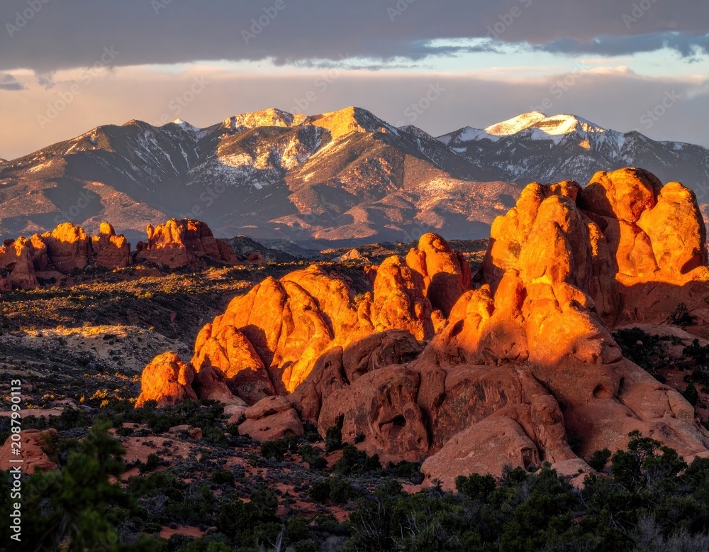 Naklejka premium Dramatic sunset illuminates sandstone formations with snow-capped mountains in the background.