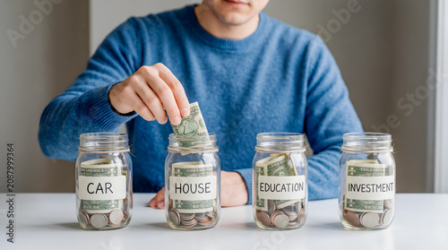 Man placing dollar bill into savings jars labeled car house education investment