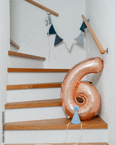 number six balloon floating on staircase interior with wooden steps and handrails, celebratory scene for birthday party home decoration, side view of staircase with blue party hat and flags.