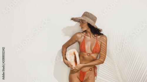 Stylish Woman In Bikini And Hat Holding Shell Against White Wall