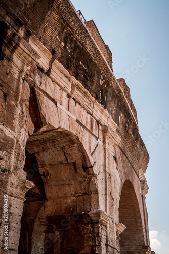 Low-angle photograph of the Colosseum exterior in Rome showing weathered stone arches and architectural details against a clear blue sky