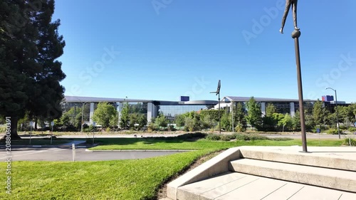 Wide panning shot of the Google campus headquarters building with its modern glass facade, surrounded by a lush green park and sculptures on a sunny day in Palo Alto, California