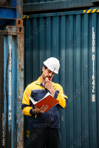 A hard-hat worker reviews documents beside stacked shipping containers, planning logistics and safety checks at a freight yard.