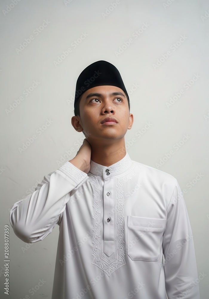 Obraz premium Young Muslim Man in Traditional White Baju Melayu and Songkok Looking Up Thoughtfully Against White Background