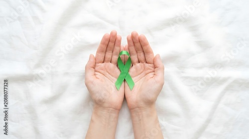 A pair of hands gently cradling a green awareness ribbon on a white background, symbolizing support and hope for mental health, wellness, and cancer causes.